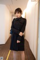 A woman in a black trench coat standing in a hallway.