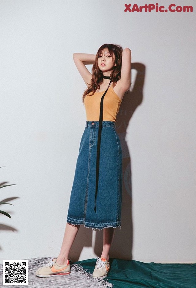 a woman standing in front of a white wall wearing a yellow top and blue denim skirt