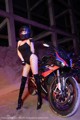 A woman in a black bodysuit and fishnet stockings standing next to a motorcycle.