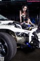 A woman standing next to a wrecked car in a garage.